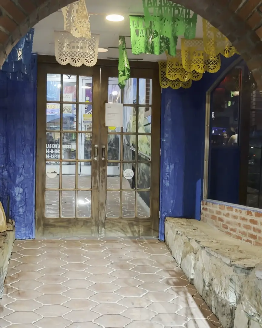 The brick arch entrance at night, lit by two lanterns, with papel picado hanging over the walkway to the front doors